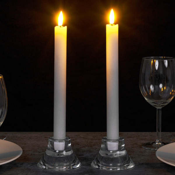 Două lumânări albe LED cu lumină caldă pâlpâitoare, aranjate pe o masă cu pahare de vin.